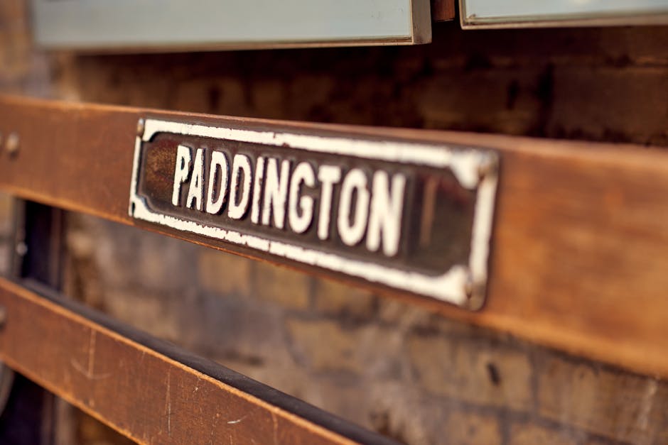 Close-up view of a vintage metal street nameplate with black background and white lettering displaying 'Paddington,' mounted on a wooden structure with a distressed finish. The wooden framework is part of a building exterior or large furniture piece, featuring visible grain and weathered textures. In the background, blurred elements suggest an indoor or outdoor environment related to house removals or furniture transportation, with soft natural lighting highlighting the aged materials. This image capturing historic street signage and wooden textures supports content about home relocation, furniture transport, or packing and moving services provided by Movers Paddington, illustrating the process of moving traditional or preserved features during a house removal on Bayswater Road.