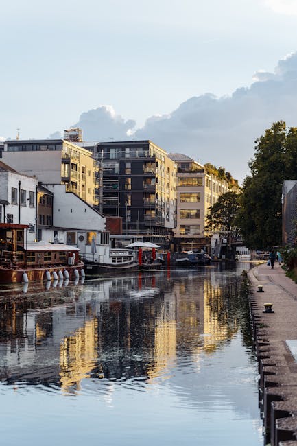 A waterway flanked by a paved walkway on the right side, with a row of modern multi-story apartment buildings and houseboats lining the far bank. The buildings feature large windows and balconies, with some displaying signage and outdoor furniture. On the water, several boats and floating structures are moored, with reflections visible on the calm surface. The sky above is partly cloudy with patches of blue, and a few trees are visible on the right side, adding greenery to the urban scene. This image captures an urban residential area potentially involved in house removals or furniture transport, relevant to the logistics of home relocation and moving services offered by Movers Paddington.