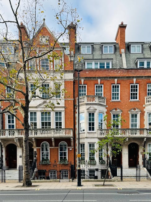 Image of a row of historic townhouses on Cornwall Terrace in W2, Paddington, with ornate brick facades, large sash windows, and decorative balconies. In the foreground, a leafless tree partially obscures the buildings, and a black metal fence runs along the pavement. The scene shows a sunny day with a partly cloudy sky. No moving activity is visible, but the setting suggests preparation for home relocation or furniture transport, with the buildings providing a backdrop for professional removals services by Movers Paddington as part of a house move focused on protecting period features.