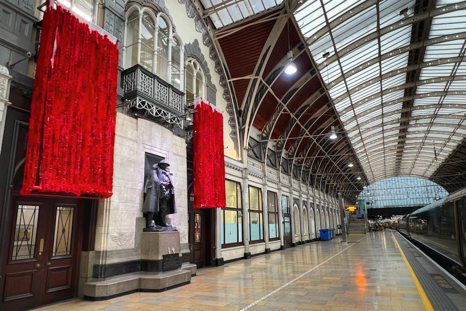 Inside a historic train station with a high arched glass and metal roof, the platform is empty with a row of classical-style windows and ornate ironwork on the facade. Two large red floral arrangements are mounted on either side of a bronze statue of a person dressed in period clothing, positioned on a stone pedestal against a stone wall. The platform surface is wooden and tiled, illuminated by overhead lighting. On the right, a modern train with sleek metallic surfaces is parked, and there are some blue recycling bins visible along the platform. The station’s spacious interior is well-lit and clean, providing a setting suitable for a home relocation or furniture transport, with visible elements of loading and moving logistics carried out by professional removals experts such as Movers Paddington.