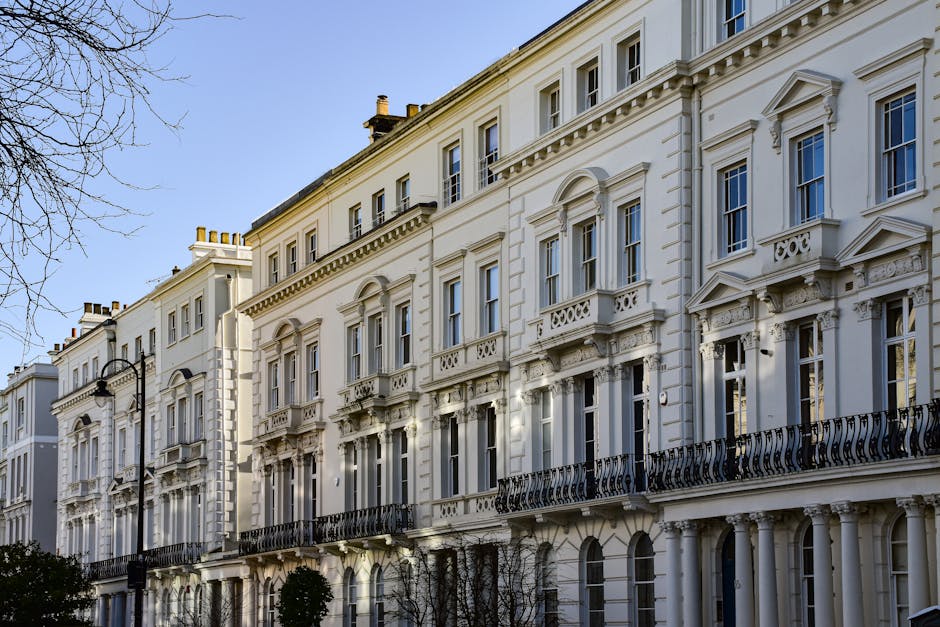 Image of a row of historic townhouses on Cornwall Terrace in W2, Paddington, with ornate brick facades, large sash windows, and decorative balconies. In the foreground, a leafless tree partially obscures the buildings, and a black metal fence runs along the pavement. The scene shows a sunny day with a partly cloudy sky. No moving activity is visible, but the setting suggests preparation for home relocation or furniture transport, with the buildings providing a backdrop for professional removals services by Movers Paddington as part of a house move focused on protecting period features.
