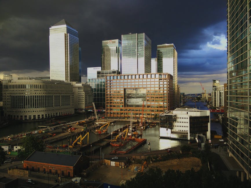 A daytime view of the Paddington Basin area, featuring a construction site with several yellow crane machines and temporary fencing along the edge of a waterway. In the foreground, there are various construction materials and equipment, including wooden pallets and metal scaffolding. Surrounding the site are modern office buildings and high-rise structures, with reflective glass facades, some with distinctive grid patterns. The sky above shows dark, stormy clouds with patches of sunlight breaking through, casting shadows across the skyline. This scene captures an active commercial environment frequently involved in property development and office relocations, which could be relevant to house removals, furniture transport, or packing and moving processes. Movements of construction vehicles and materials suggest ongoing work, potentially in preparation for a residential or business building transition, aligned with professional relocation services by [COMPANY_NAME].