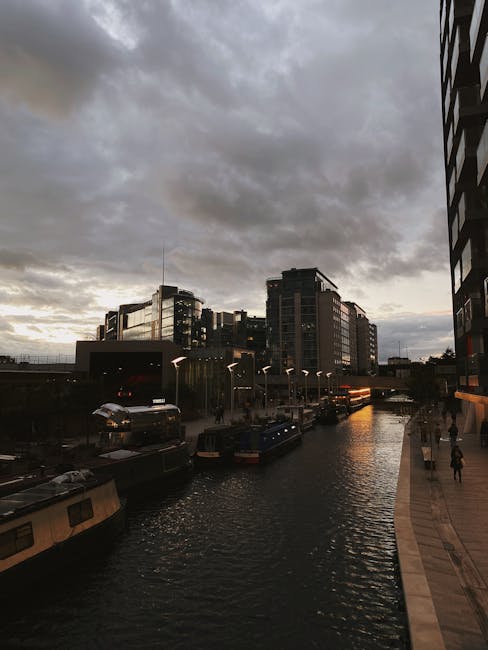 Image showing a scene of a canal or waterway in Paddington with several houseboats moored along the edge, visible from an elevated perspective during late afternoon or early evening with overcast skies. Modern office buildings and residential structures with reflective glass windows are situated behind the water, some featuring balconies. A narrow walkway runs parallel to the water, with a few pedestrians walking along it, and streetlights are illuminated, creating a warm glow against the cloudy sky. The environment suggests an urban setting with a combination of commercial and residential buildings near the water. The image captures the atmosphere of a busy city area, potentially during a house removal or relocation process, where the scenic urban landscape provides a backdrop for moving activities coordinated by companies like Movers Paddington as part of a household or office relocation project near Paddington Basin. The lighting indicates natural daylight fading into dusk, emphasizing the transition from day to night in this cityscape.