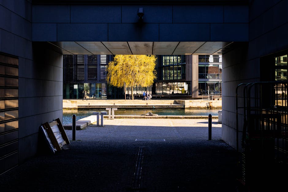 A daytime view of the Paddington Basin area, featuring a construction site with several yellow crane machines and temporary fencing along the edge of a waterway. In the foreground, there are various construction materials and equipment, including wooden pallets and metal scaffolding. Surrounding the site are modern office buildings and high-rise structures, with reflective glass facades, some with distinctive grid patterns. The sky above shows dark, stormy clouds with patches of sunlight breaking through, casting shadows across the skyline. This scene captures an active commercial environment frequently involved in property development and office relocations, which could be relevant to house removals, furniture transport, or packing and moving processes. Movements of construction vehicles and materials suggest ongoing work, potentially in preparation for a residential or business building transition, aligned with professional relocation services by [COMPANY_NAME].