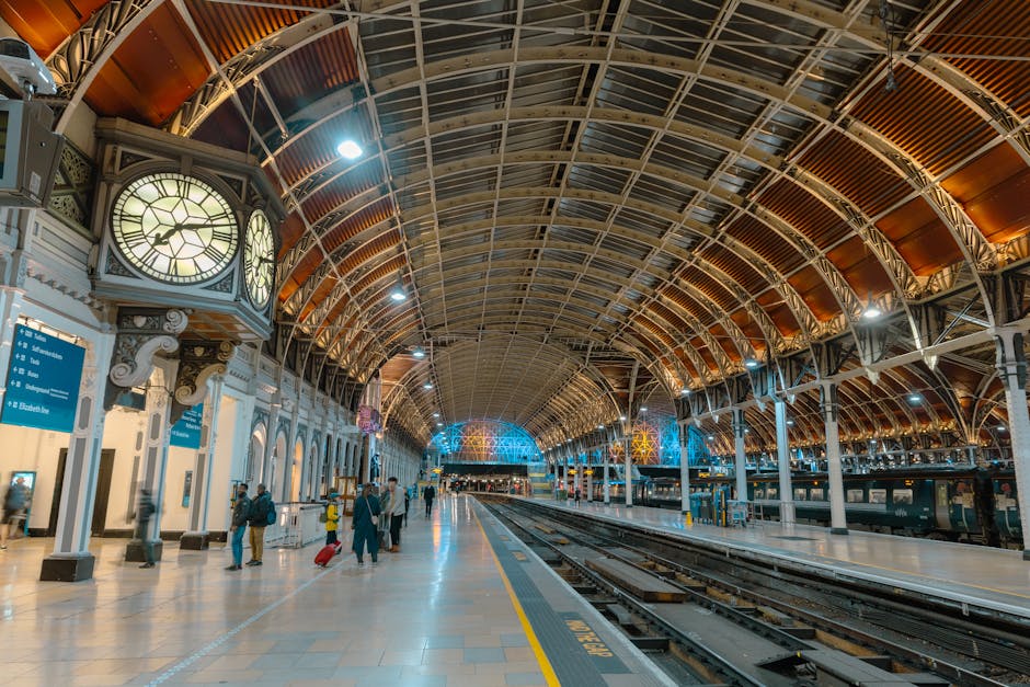 Inside a spacious train station with a high, arched metal and glass ceiling, illuminated by ceiling lights and colorful decorative window panels. On the left side, there are white-painted columns and a large ornate clock display featuring two clock faces, with people walking along the platform. Some passengers are carrying luggage or pulling wheeled suitcases, preparing for departure or arrival. The platform is clean and well-lit, with train tracks running parallel along the right side, and train carriages are visible at the far end of the platform. The environment is calm, with a mix of standing and walking individuals, and equipment such as luggage trolleys and platform markings are evident. This scene showcases the logistics of home relocation or furniture transport within a busy transportation hub, with Movers Paddington providing insights into packing and moving processes related to house removals and freight handling.