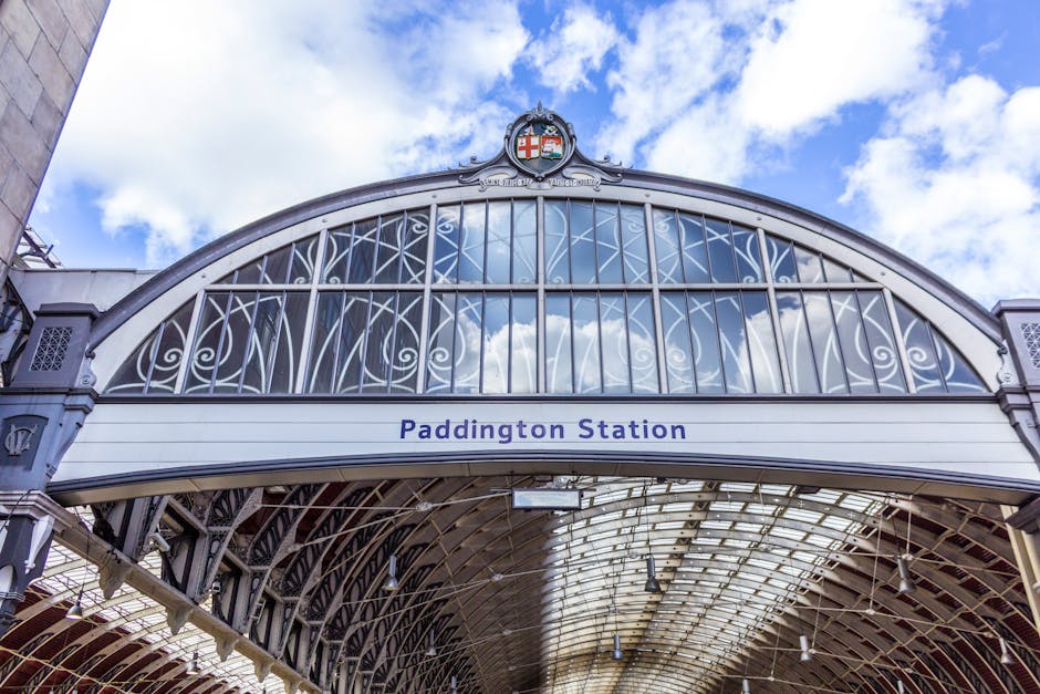 Exterior view of Paddington Station showcasing a curved, ornate metal and glass roof structure with intricate white metalwork framing the glass panels and a coat of arms emblem at the peak. The station's name, 'Paddington Station,' is printed in blue on a white horizontal sign below the arch. The bright sky with scattered clouds is reflected in the glass, and part of the station's stone and metal facade is visible on the left side. This architectural detail highlights the historic design elements typical of railway stations involved in home relocation and furniture transport services, supported by providers like Movers Paddington, who assist with packing, loading, and transportation logistics.