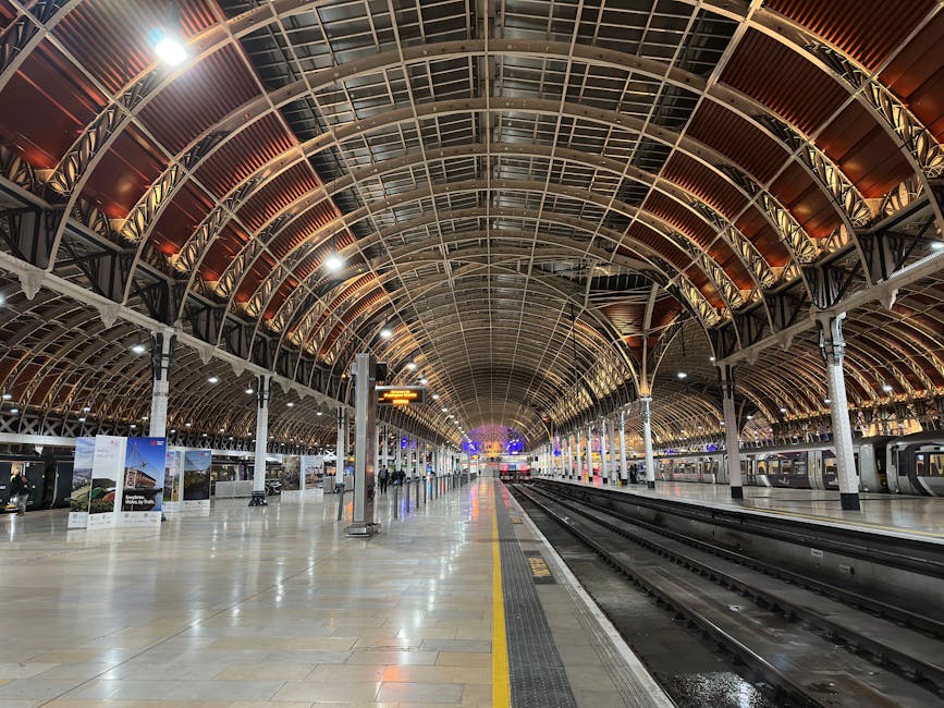 Inside a spacious train station with a high, arched metal and glass ceiling, illuminated by ceiling lights and colorful decorative window panels. On the left side, there are white-painted columns and a large ornate clock display featuring two clock faces, with people walking along the platform. Some passengers are carrying luggage or pulling wheeled suitcases, preparing for departure or arrival. The platform is clean and well-lit, with train tracks running parallel along the right side, and train carriages are visible at the far end of the platform. The environment is calm, with a mix of standing and walking individuals, and equipment such as luggage trolleys and platform markings are evident. This scene showcases the logistics of home relocation or furniture transport within a busy transportation hub, with Movers Paddington providing insights into packing and moving processes related to house removals and freight handling.