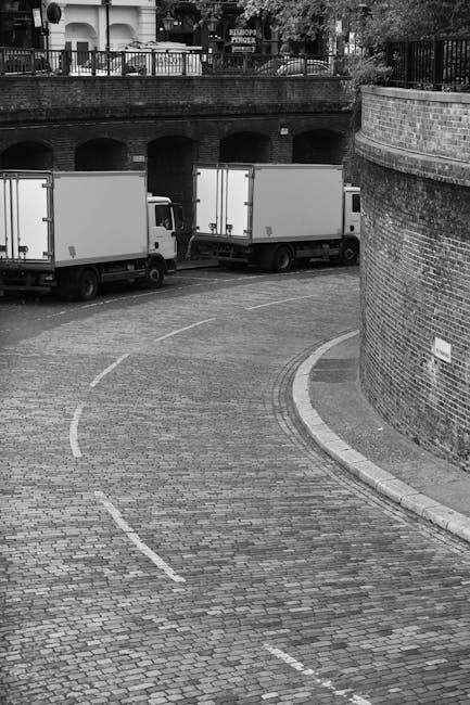 Black and white photograph of a narrow, curved cobblestone street bordered by a brick wall on the right side and a metal railing above it, with two white moving trucks parked along the curb. The trucks are positioned parallel to the wall, with the rear doors facing the street, indicating they are in the process of loading or unloading. In the background, there is an arched brick underpass beneath the railing, with some residential or commercial buildings visible above, including windows and trees. The scene illustrates a home relocation or furniture transport operation, with clean, well-organized vehicles used by Movers Paddington, set against the urban environment typical of Paddington, W2, during daylight hours.