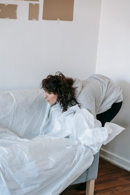 A woman with dark, curly hair wearing a grey hoodie is inside a room, leaning over a sofa wrapped in white packing paper or moving blankets, which is likely being prepared for a house relocation. The sofa is positioned against a white wall with three rectangular patches or labels attached to it, possibly indicating parts to be disassembled or areas for marking during packing. The room has a wooden floor, and natural lighting illuminates the scene. This image illustrates the process of packing or preparing furniture for a move, relevant to home removals and furniture transport services provided by Movers Paddington, as part of the house or flat removal process described in the webpage about small move tips for a studio flat on Sussex Gardens.