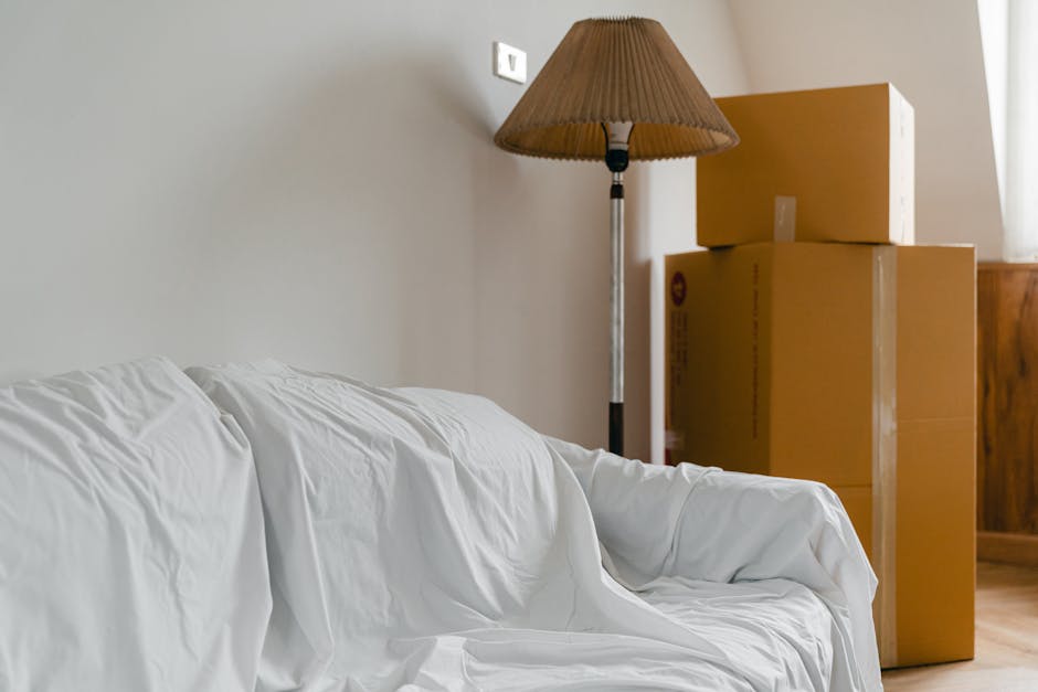 Inside a room during a house move, four brown cardboard moving boxes are stacked on the right side of the image next to a wooden furniture piece. The boxes are sealed with clear packing tape and vary in size, with the larger one at the bottom. To the left in the foreground, a white fabric sofa with visible creases and slightly wrinkled upholstery is partially visible. Behind the boxes, there is a tall floor lamp with a brown pleated lampshade, positioned against a plain, light-colored wall. The room is well-lit by natural light coming from a window on the right, which is partially visible. A white electrical socket is mounted high on the wall, and the overall scene depicts a home during the process of packing and moving, with furnishings and packing materials indicating a transitioning phase of a home relocation. The environment and the visible items suggest preparations involved in furniture transport and house removals, with the setting aligned to house relocation services provided by Movers Paddington.