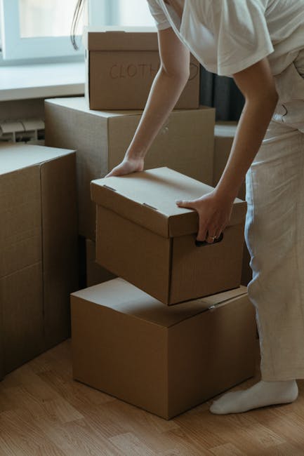 A woman with dark, curly hair wearing a grey hoodie is inside a room, leaning over a sofa wrapped in white packing paper or moving blankets, which is likely being prepared for a house relocation. The sofa is positioned against a white wall with three rectangular patches or labels attached to it, possibly indicating parts to be disassembled or areas for marking during packing. The room has a wooden floor, and natural lighting illuminates the scene. This image illustrates the process of packing or preparing furniture for a move, relevant to home removals and furniture transport services provided by Movers Paddington, as part of the house or flat removal process described in the webpage about small move tips for a studio flat on Sussex Gardens.