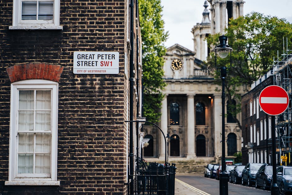 A two-storey brick building on Great Peter Street in Westminster, London, with a white-framed sash window on the ground floor and a small sign indicating the street name and postcode. Behind the building, a historic stone church with columns and a clock face is visible, partially obscured by trees with green leaves. To the right, there is a black lamppost and a red and white no-entry traffic sign on the street corner. Parked cars line the street, and a construction scaffold is present on the right side, indicating ongoing building work. The scene is illuminated by natural daylight, suggesting a clear day, and the overall setting illustrates an urban environment suitable for professional house removals and furniture transport, with festival and moving logistics potentially involved.