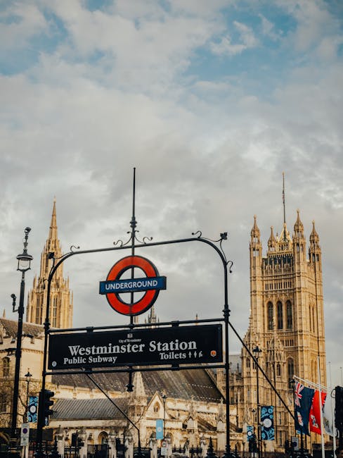 A street view showing the entrance to Westminster Station in London, with a prominent London Underground roundel sign featuring a red circle and blue bar that says 'UNDERGROUND'. Below the sign is a black informational panel indicating 'Westminster Station' along with additional details about public subway services and toilets. In the background, the historic Gothic-style towers of the Palace of Westminster are visible, with intricate stonework and spires reaching into a partly cloudy sky. The scene is set during daylight hours, and the area around the station appears to be a busy urban environment, often associated with house removals or home relocation activities in Westminster, where careful planning for furniture transport and loading processes is essential. The image reflects an iconic London setting relevant to moving services such as those offered by Movers Paddington, particularly during packing and loading phases of a house move near central London landmarks.