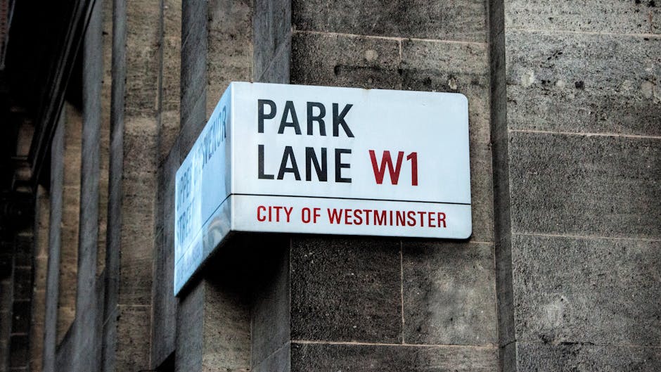 A close-up view of a rectangular white street sign mounted on a brick building wall, displaying the words 'PARK LANE W1' in bold black and red text. Beneath, in smaller red letters, it reads 'CITY OF WESTMINSTER.' The sign is attached to the corner of the building, adjacent to a metal gate or fencing structure. Visible in the background are part of the building's textured stone or brick surface and nearby architectural elements. This urban scene relates to street naming and parking zone designations within Westminster, relevant to house removals and moving logistics in the Paddington area, as managed or referenced by Movers Paddington.